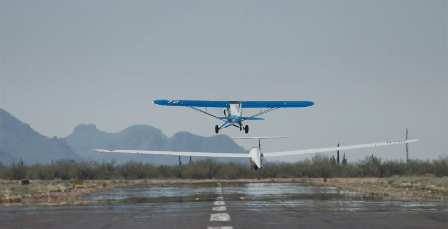 One silver and one blue plane take off from a runway, with clear skies and mountains in the distance.